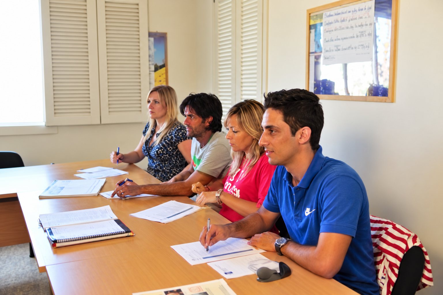 Quatre personnes, deux femmes et deux hommes, sont assises côte à côte à une table couverte de documents dans une salle aux volets blancs.